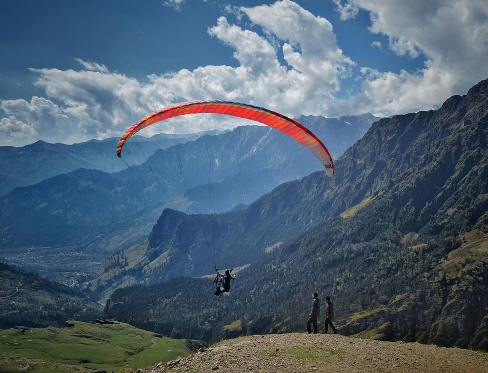 Paraglider with an orange wing flying over a wide green valley at Bir Billing